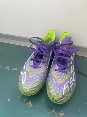 Adidas F50 lila/gula fotbollsskor  - Säljer ett par Adidas F50 fotbollsskor i en riktigt fet färgkombo av lila, vitt och neon-gult. Skorna har snörning, låg profil och mönstrad ovandel i syntetmaterial för max speed på planen. Sulan har fasta dobbar för gräs. Perfekta för dig som vill sticka ut på matchen!