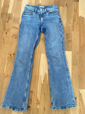 Blå bootcut jeans med låg midja - Snygga blå bootcut jeans med låg midja och klassiska fem fickor. Jeansen har en lätt tvättad look och vida ben som ger en retrovibe. Perfekta för dig som gillar en avslappnad men trendig stil. Materialet är denim i bomull.