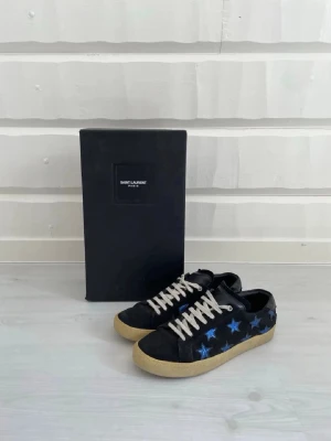 Saint Laurent sneakers med blå stjärnor - Snygga svarta Saint Laurent sneakers med blå stjärnor på sidorna och vita skosnören. Skorna har en klassisk låg siluett och beige sula. Materialet är troligtvis läder eller mocka, vilket ger en exklusiv känsla. Perfekta för dig som vill sticka ut med coola detaljer.pris kan diskuteras. Har tappat lite färg på stjärnorna 
