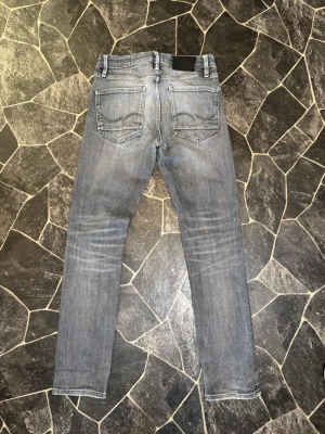 Slim Straight Tim jeans från JacknJones - Säljer ett par grå slim fit jeans med klassisk femficksmodell och diskreta slitningar. Jeansen har normal midja och raka ben. Perfekta för en avslappnad och trendig look.