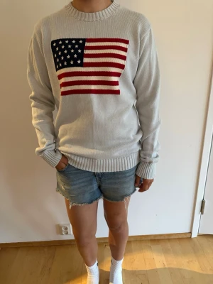 Ljusblå stickad tröja Brandy Melville - Ljusblå stickad tröja från Brandy Melville med amerikansk flagga på bröstet. Tröjan har rund halsringning och ribbade muddar vid ärmslut och nederkant. Perfekt för en avslappnad och cool stil.