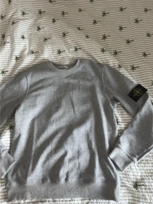 Grå sweatshirt från Stone Island - Säljer en stilren grå sweatshirt från Stone Island med den klassiska svarta patchen med gult och grönt märke på vänster ärm. Tröjan har rund halsringning, ribbade muddar och är gjord i mjukt bomullsmaterial. Perfekt för en avslappnad streetwear-look.