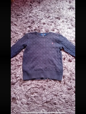 Mörkblå kabelstickad tröja Polo Ralph Lauren - Snygg mörkblå kabelstickad tröja från Polo Ralph Lauren med rund halsringning och diskret röd logga broderad på bröstet. Tröjan har ribbade muddar vid ärmslut och nederkant. Perfekt för lager på lager och klassisk stil.