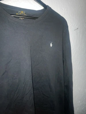 Svart långärmad tröja Polo Ralph Lauren - Klassisk svart långärmad tröja från Polo Ralph Lauren med rund halsringning och den ikoniska vita logotypen broderad på bröstet. Tillverkad i mjuk bomull som känns skön mot huden. Perfekt till jeans eller chinos för en clean look.   Urtvättad och print vid kragen har börjat lossna!  Fick denna i födelsedagspresent!  Hör av dig vid mer frågor!  