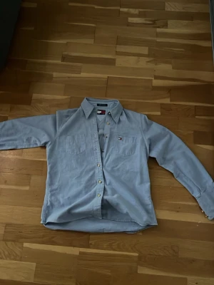 Ljusblå skjorta från Tommy Hilfiger - Klassisk ljusblå skjorta från Tommy Hilfiger i 100% bomull. Skjortan har lång ärm, knappar framtill och två bröstfickor. Ikonisk logga på vänster bröstficka. Perfekt för en clean och stilren look.