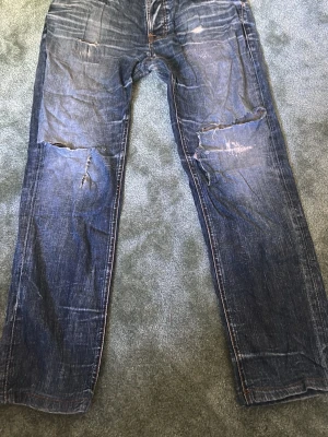 Blå slitna jeans från Nudie Jeans - Ett par blå jeans från Nudie Jeans med tydliga slitningar och hål på benen. Klassisk femficksmodell med orangea sömmar och knappgylf. Jeansen har en rak passform och är tillverkade i denim av bomull.