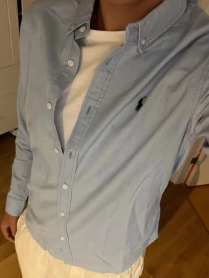 Ljusblå skjorta från Ralph Lauren - Klassisk ljusblå skjorta från Ralph Lauren med button-down krage och den ikoniska loggan broderad på bröstet. Skjortan har långa ärmar, vita knappar och är tillverkad i mjuk bomull. 