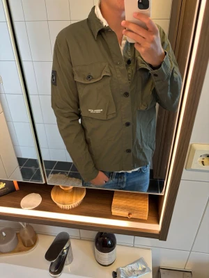 Olivgrön overshirt från Marshall Artist - Snygg olivgrön overshirt från Marshall Artist med två bröstfickor, knappstängning och patch på ärmen. Tillverkad i slitstark bomull med modern passform och diskreta logodetaljer. Perfekt lager på lager-plagg för en avslappnad streetwear-look.