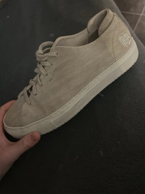 Beige sneakers från KAVAT i tyg - Snygga beige sneakers från KAVAT i canvasmaterial med vita sulor och klassisk låg profil. Skorna har snörning framtill och en diskret logga på sidan samt ett broderat märke på hälen. Perfekta för en clean och stilren look.