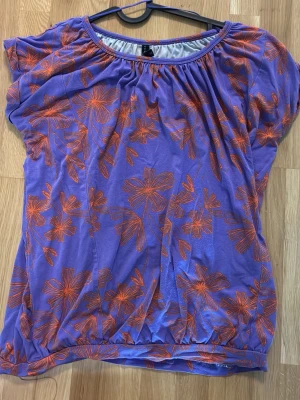 Lila blommig blus med orange mönster - Snygg lila blus med orange blommigt mönster och rund halsringning. Modellen är lös och har korta ärmar med lätt puff. Perfekt för dig som gillar färg och vill sticka ut lite extra. Tillverkad i mjuk lyocell och elastan som känns skön mot huden.