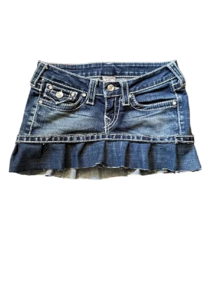 Blå jeanskjol med volang från True Religion - Säljer en blå jeanskjol från True Religion med volangkant nedtill och dekorativa fickor både fram och bak. Jättefin och cool nu på sommaren. Midjemått 35 cm.