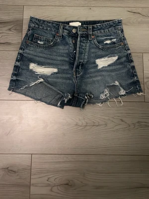 Högmidjade blå jeansshorts - Snygga högmidjade jeansshorts i blå denim med slitna detaljer och fransig kant. Klassisk femficksmodell med knappar och dragkedja framtill. Perfekta för sommarens alla äventyr.