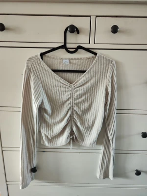 Beige ribbad croppad topp med snörning - Trendig beige croppad topp med ribbad struktur och v-ringning. Toppen har långa ärmar och snygg snörning framtill som ger en rynkad effekt. Perfekt till jeans eller kjol för en cool vardagslook. Den är i barn storlek alltså då borde de passar de runt 13 år