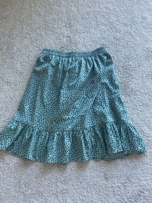 Grön prickig omlottkjol med volang - Söt grön kortkjol med svarta prickar och volangkant. Kjolen har omlottmodell med knytband i midjan och är gjord i ett lätt, flowy material som passar perfekt till varma dagar. Perfekt för dig som gillar en lekfull och trendig stil.