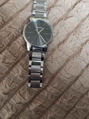 Silverfärgad klocka från Calvin Klein - Stilren klocka från Calvin Klein med rund svart urtavla och silverfärgat armband i metall. Minimalistisk design med diskreta markeringar och klassiskt spänne. Perfekt accessoar för dig som gillar enkel och modern stil.