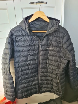 Svart dunjacka med huva från Peak Performance - Snygg svart dunjacka från Peak Performance med huva och dragkedja framtill. Den är lite liten i storlek så passar nog någon med storlek L perfekt. Jackan har en quiltad design, två sidofickor med dragkedja och diskret logga på bröstet. Perfekt för att köra moped på våren eller användas på hösten/vintern. Den är inte särskilt tjockt vadderad men håller värme bra och ventilerar samtidigt😊 