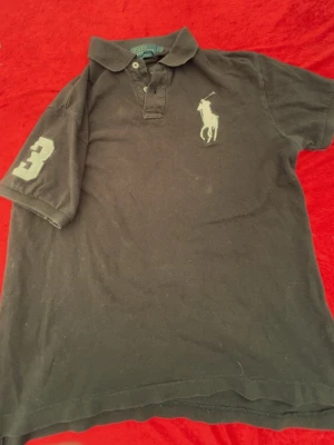 Svart pikétröja från Ralph Lauren - Klassisk svart pikétröja från Ralph Lauren med vit broderad logga på bröstet och en stor siffra 3 på ärmen. Tröjan har krage, knappar vid halsen och är kortärmad. Perfekt för en sportig och stilren look.