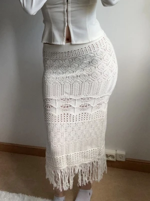 Vit virkad kjol - Supersnygg vit långkjol i virkad stil med olika mönster och dekorativa fransar längst ner. Kjolen har en kroppsnära passform och elastisk midja, perfekt för en trendig och bohemisk look.