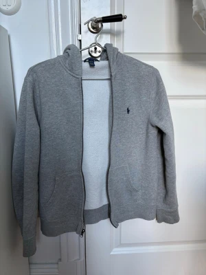 Grå hoodie från Polo Ralph Lauren - Klassisk grå hoodie från Polo Ralph Lauren med dragkedja framtill och två fickor. Liten mörkblå logga broderad på bröstet. Tillverkad i mjuk bomullsblandning, perfekt för chill dagar. Enkel och stilren design som funkar till det mesta. Äkta - inköpt 2025.