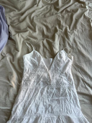 Vit sommartopp med spetsdetaljer - Söt vit topp/klänning med tunna axelband och v-ringning. Den har fina spetsdetaljer och broderier framtill samt längs kjolen. Perfekt för varma dagar och har en lätt, luftig känsla.