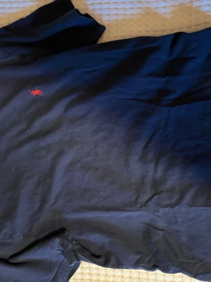 Mörkblå t-shirt från Polo Ralph Lauren - Snygg mörkblå t-shirt från Polo Ralph Lauren med klassisk röd logga broderad på bröstet. T-shirten är kortärmad och tillverkad i mjuk bomull, perfekt för en stilren och avslappnad look. Den passar för barn med strl M. 
