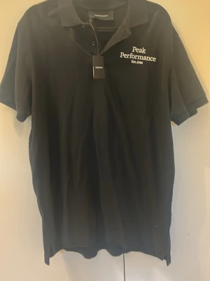 Svart Peak Performance pikétröja - Svart pikétröja från Peak Performance med vit broderad logga på bröstet. Klassisk krage, knappar framtill och korta ärmar. Tillverkad i mjuk bomull för en bekväm och stilren look.