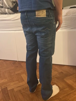 Blå jeans från Jacob Cohen - Säljer ett par snygga blå jeans från Jacob Cohen med klassisk femficksmodell och raka ben. Jeansen har diskreta slitningar och kontrastsömmar samt en beige patch med logga bak i midjan. Tillverkade i mjukt denimtyg för skön passform.