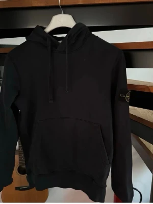 Svart hoodie från Stone Island - Säljer en svart hoodie från Stone Island med klassisk patch på vänster ärm. Hoodien har huva med dragsko och en stor magficka framtill. Tillverkad i mjukt bomullsmaterial och har ribbade muddar vid ärmslut och nederkant. Perfekt för en avslappnad och stilren look.