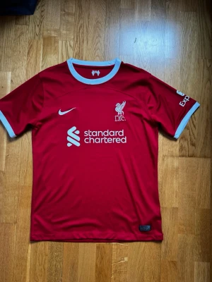Liverpool matchtröja Nike röd - Säljer en röd Liverpool FC matchtröja från Nike med vita detaljer vid krage och ärmslut. Tröjan har klubbmärke och Nike-logga på bröstet samt sponsortryck framtill. Tillverkad i lätt och ventilerande funktionsmaterial, perfekt för fotbollsträning.