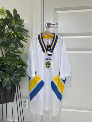Svensk landslagströja Adidas - Vit svensk landslagströja från Adidas med blå och gula detaljer, samt broderad svensk sköld på bröstet. Tröjan har v-ringad krage med blå och gula ränder och är tillverkad i lätt, ventilerande material. Perfekt för fotboll eller supporterhäng.