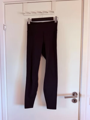 Svarta träningstights från H&M - Svarta, långa träningstights från H&M med hög midja och tight passform. Perfekta för gym, löpning eller yoga. Materialet är stretchigt och följsamt för maximal rörelsefrihet. Diskret logga på ena benet. Sparsamt använda