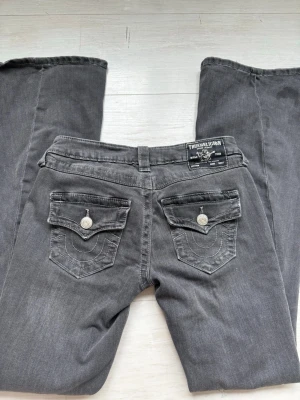 Grå bootcut jeans True Religion - Säljer ett par gråa bootcut jeans från True Religion med ikoniska ficklock och dekorativa sömmar bak. Jeansen har låg midja, silverfärgade knappar och är tillverkade i ett mjukt denimtyg. Perfekta för en avslappnad men snygg look. Använt fåtal gånger