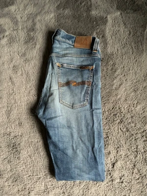Blå Nudie Jeans med slitningar - Säljer ett par blå jeans från Nudie Jeans grim Tim med slitningar och hål på benen för en cool, avslappnad look. Storlek 28/30 men sitter mer som 30/30. Ett litet hål som syns på bild 3 men borde lätt gå att fixa eller sy ihop. 