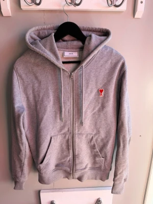 Grå hoodie med dragkedja från Ami - Snygg grå hoodie från Ami med dragkedja framtill och två stora fickor. På bröstet sitter Ami:s ikoniska röda hjärta med bokstaven A. Tillverkad i mjuk bomullsblandning och har justerbar huva med snören. Perfekt för en avslappnad och trendig look.