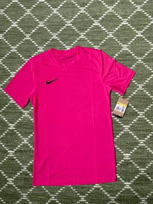 Rosa Nike Dri-FIT träningströja - Snygg rosa träningströja från Nike med svart swoosh-logga på bröstet. Tillverkad i Dri-FIT-material som transporterar bort fukt och håller dig torr under träningen. Klassisk rund halsringning och korta ärmar, perfekt för gymmet eller löprundan.