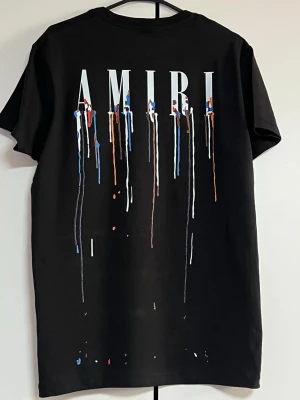 Svart AMIRI t-shirt med färgstänk - Cool svart t-shirt från AMIRI med stort tryck på ryggen där märkesnamnet syns i vitt och färgstänk i blått, rött, orange och vitt som rinner ner. T-shirten har klassisk passform och är kortärmad, perfekt för dig som gillar streetwear och statement-plagg.