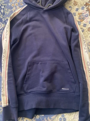 Blå hoodie från Gant med logga - Snygg blå hoodie från Gant med huva och magficka. Ärmarna har vita paneler med röda linjer och GANT-logga tryckt längs hela armen. Tillverkad i mjukt bomullsmaterial som känns skönt mot huden. Perfekt för en avslappnad och sportig stil.