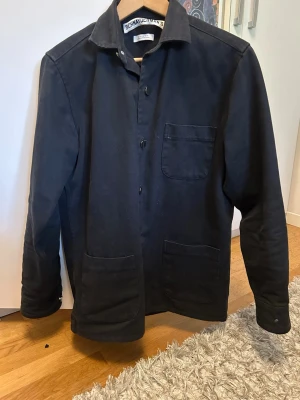 Svart overshirt från Schnayderman's - Stilren svart overshirt från Schnayderman's med klassisk krage och tre framfickor. Rak passform och knäppning framtill. Perfekt lager-på-lager-plagg i ett slitstarkt bomullstyg.