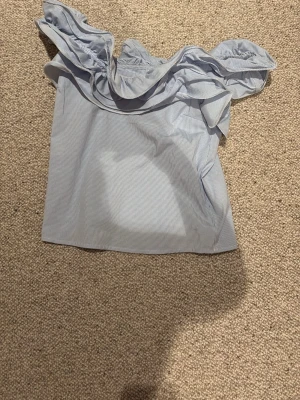 Blå offshoulder topp med volang - Söt blå offshoulder topp med tunna vita ränder och bred volang runt axlarna. Perfekt för dig som gillar en luftig och trendig stil. Toppen är ärmlös och har en croppad passform som passar till både jeans och kjol.