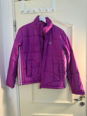 Lila pufferjacka från Adidas - Säljer en lila pufferjacka från Adidas med klassiska vita ränder på sidorna och Adidas-logga på bröstet. Jackan har hög krage, dragkedja framtill och två stora fickor. Perfekt för kalla dagar och riktigt snygg färg som sticker ut.