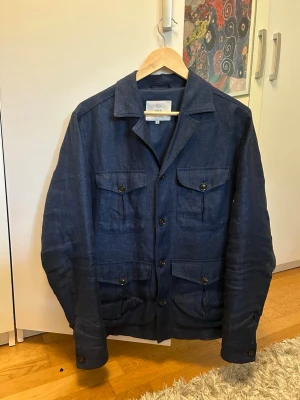 Mörkblå overshirt från Bläck - Snygg mörkblå overshirt från Bläck i linne. Klassisk design med fyra fickor framtill och knappar längs hela fronten. Perfekt att bära som lager över t-shirt eller skjorta. Skön och luftig känsla, idealisk för vår och sommar.
