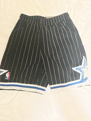 Svarta NBA shorts med vita ränder - Säljer ett par svarta NBA basketshorts med vita vertikala ränder och blå detaljer. Shortsen har elastisk midja, stora stjärnor på sidorna och en NBA-logga framtill. Perfekta för dig som gillar sportig stil och streetwear.
