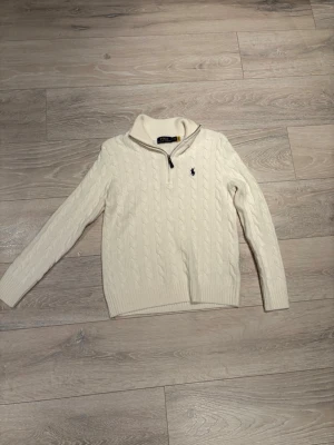 Vit kabelstickad tröja Polo Ralph Lauren - Kabelstickad vit tröja från Polo Ralph Lauren med halv dragkedja och hög krage. Tröjan har lång ärm, ribbade muddar och den klassiska blå logotypen broderad på bröstet. Perfekt för dig som gillar stilrena och tidlösa plagg.