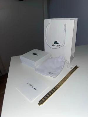 Guldigt armband från Lacoste - Snyggt guldigt armband från Lacoste med geometriskt mönster och diskret krokodil-logo. Armbandet är platt och har en elegant design som passar perfekt till din stil. Levereras med original ask, påse och presentpåse från Lacoste.