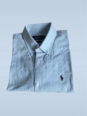 Ljusblå skjorta från Ralph Lauren - Klassisk ljusblå skjorta från Ralph Lauren med button-down krage och broderad logga på bröstet. Skjortan har lång ärm, vita knappar och är tillverkad i mjuk bomull. Perfekt för en clean och stilren look.