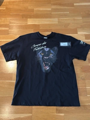 Svart Comme des Fuckdown t-shirt - Svart t-shirt från Comme des Fuckdown med stor tryckt panter på bröstet och vit text ovanför. Klassisk men lite oversized passform med ärmar och rund hals. Materialet känns som mjuk bomull och trycket ger en edgy vibe. Denna modell fraktas normalt från Italien, men jag säljer den här i Sverige → snabbare och billigare leverans! OBS! det står storlek small men den är lite oversized, så lite mer en medium. Den är helt ny då den inte passade mig, prisetiketten är kvar samt original påse. 