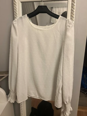 Vit blus med strukturmönster - Säljer en vit blus med diskret strukturmönster och rund halsringning. Blusen har långa ärmar med smockade muddar och en luftig passform. Perfekt för dig som gillar stilrena och klassiska plagg.