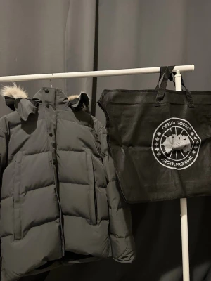 Grå Canada Goose Wyndham Jacka med Äkta päls - Snygg och varm dunjacka från Canada Goose i grå färg med äkta päls på huvan. Klassisk pufferstil med dragkedja och knappar framtill, samt avtagbar luva/päls. Ikonisk logga på ärmen och kommer med alla tillbehör.  Perfekt för kalla vinterdagar – håller värmen samtidigt som den ser exklusiv ut. Pris kan diskuteras vid snabb affär. Fraktas endast.