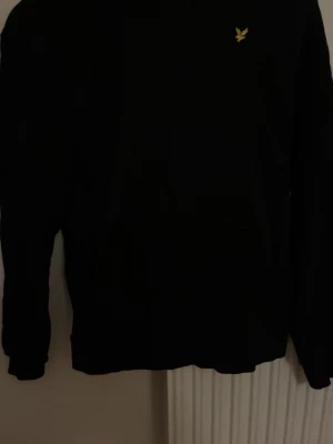 Svart sweatshirt från Lyle & Scott - Svart sweatshirt från Lyle & Scott med klassisk gul logga broderad på bröstet. Tröjan har rund halsringning, långa ärmar och ribbade muddar. Perfekt för en stilren och enkel look. Nypris ca 700kr