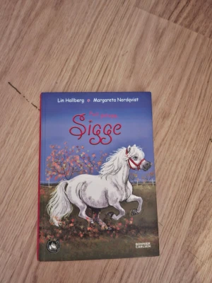 Full galopp Sigge av Lin Hallberg - Säljer boken 'Full galopp Sigge' av Lin Hallberg och Margareta Nordqvist. Boken har hård pärm med färgglad illustration av en vit ponny på framsidan och röd rygg. Perfekt för dig som gillar hästböcker och äventyr.